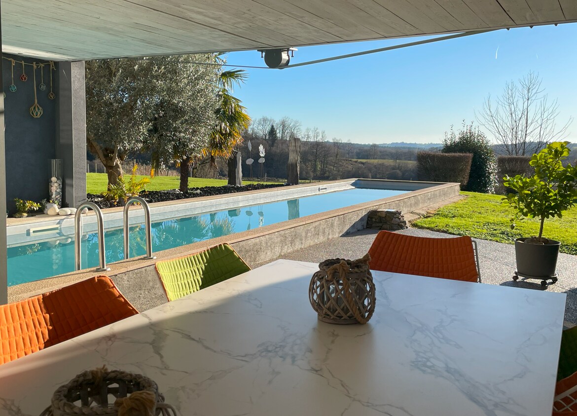 A covered outdoor terrace features a white table set with decorative accents. Brightly colored chairs in shades of orange and green surround the table. Beyond, a modern swimming pool reflects sunlight, bordered by lush greenery and expansive views of the Limousin countryside.