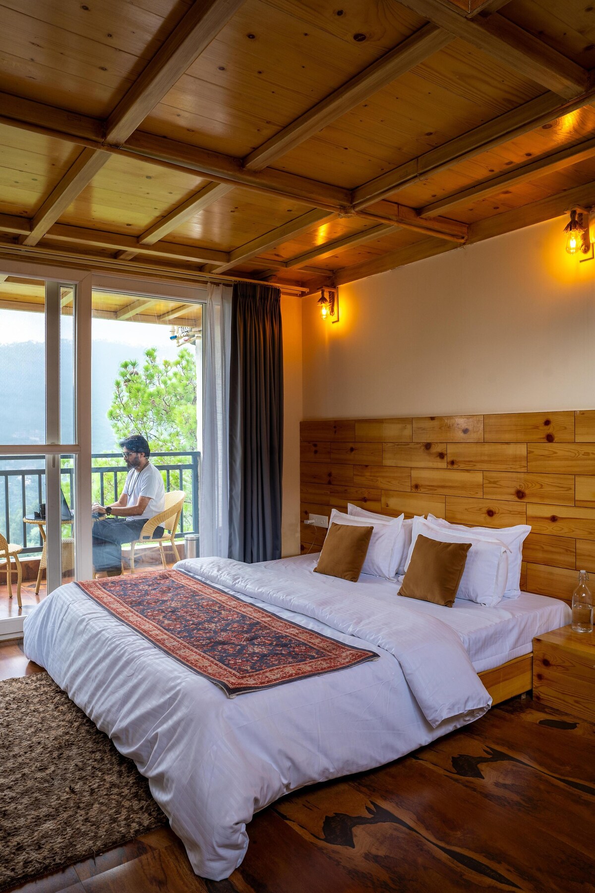 A cozy sleeping area features a spacious bed with white linens, complemented by decorative pillows. Natural wood paneling frames the room, and large glass doors open to a balcony showcasing mountain views. A soft area rug lies beneath the bed, enhancing the inviting ambiance.