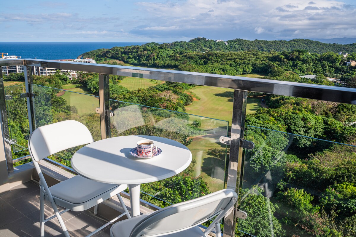 A small balcony features a round table set with a decorative bowl and two chairs, overlooking a lush landscape. The view includes rolling green hills and glimpses of the ocean in the distance, creating a serene outdoor space for relaxation.