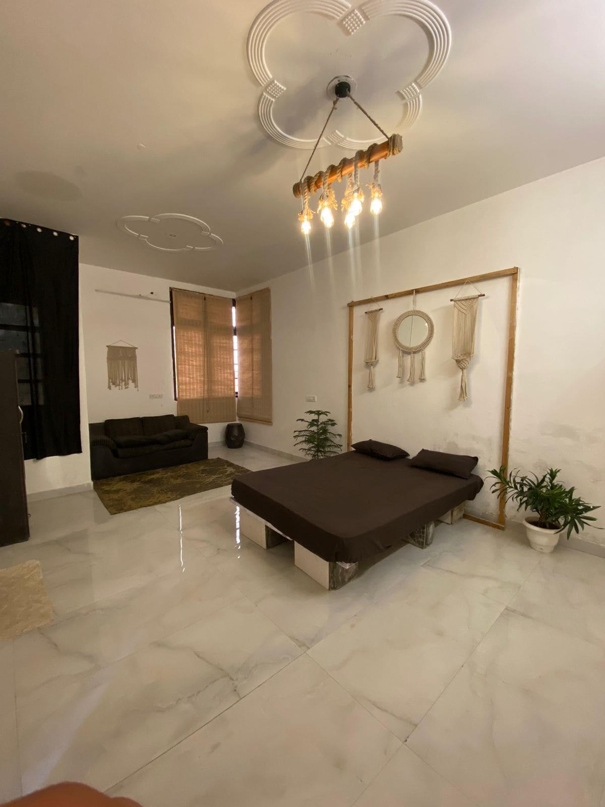 A spacious room is highlighted by glossy white tile flooring. A low, dark bed frame is centered, with matching black cushions. A cozy seating area features a dark sofa against the wall. Natural light filters through two large windows covered by light blinds.