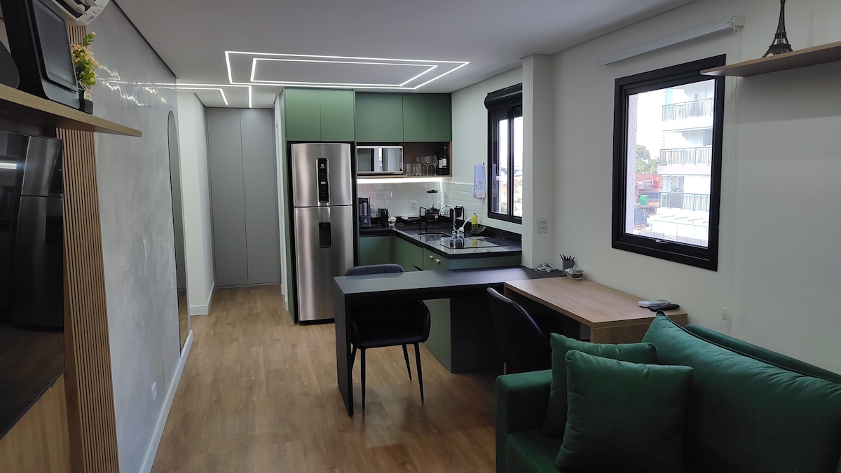 A modern kitchen and living area are presented, featuring sleek green cabinetry and stainless steel appliances. A compact dining table is set next to the kitchen. Natural light floods the space through black-framed windows, highlighting the wood flooring and comfortable green sofa.
