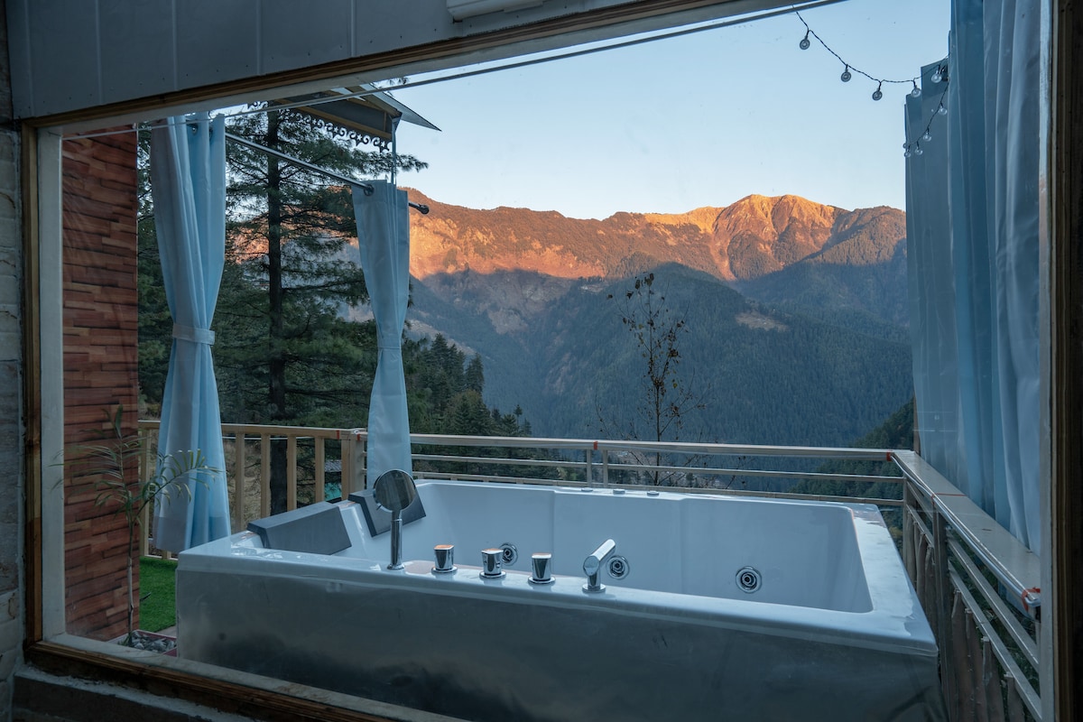 A private Jacuzzi is visible through a large glass window, offering serene views of the mountains bathed in golden light. Sheer curtains frame the window, creating a bright and airy feel, while the surrounding outdoor deck enhances the natural surroundings.