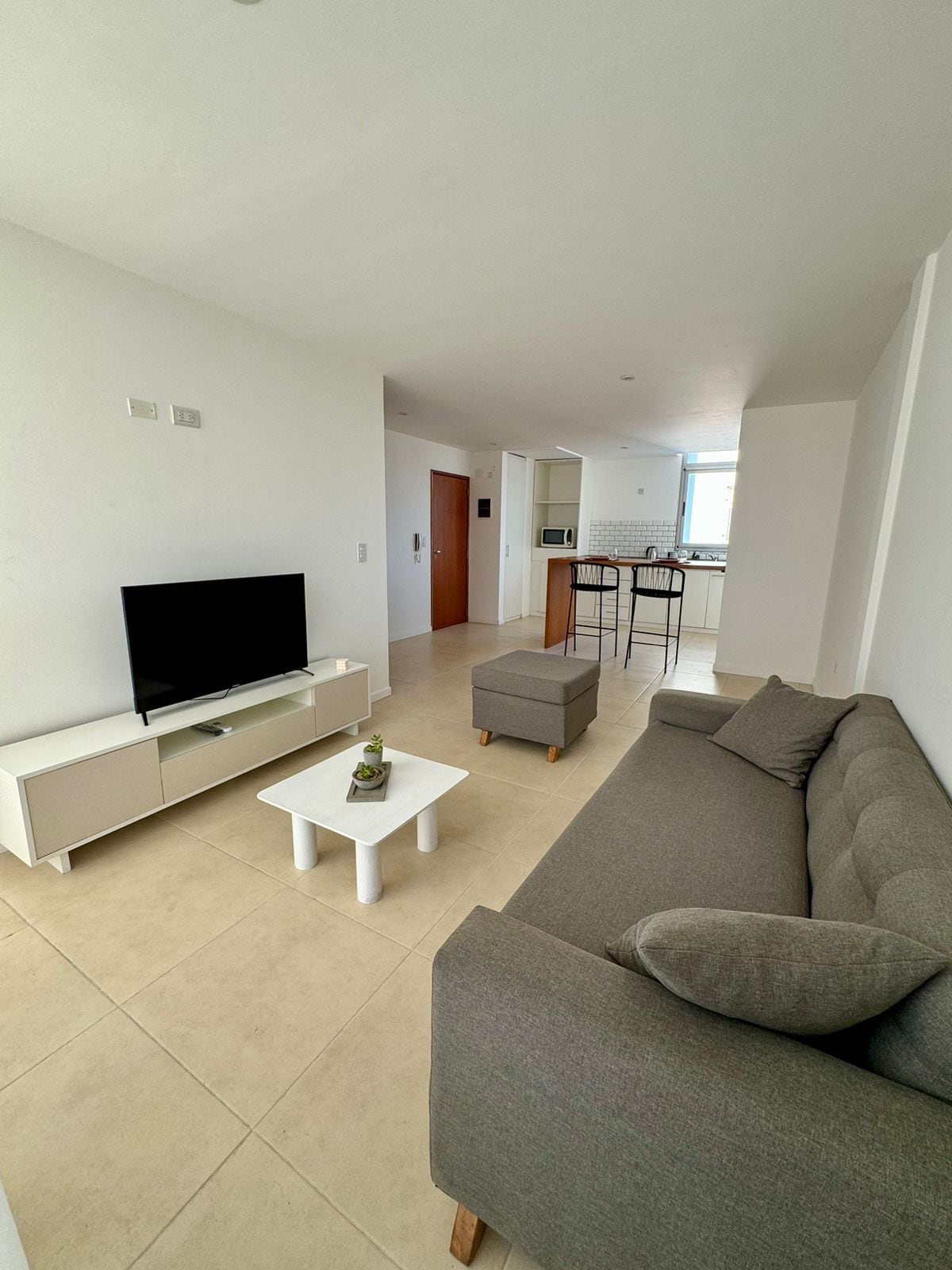 A modern living area features a comfortable gray sofa and a small ottoman. A white coffee table occupies the center space, while a flat-screen TV is positioned on a sleek console. Natural light floods in from a nearby window, enhancing the bright and airy feel.
