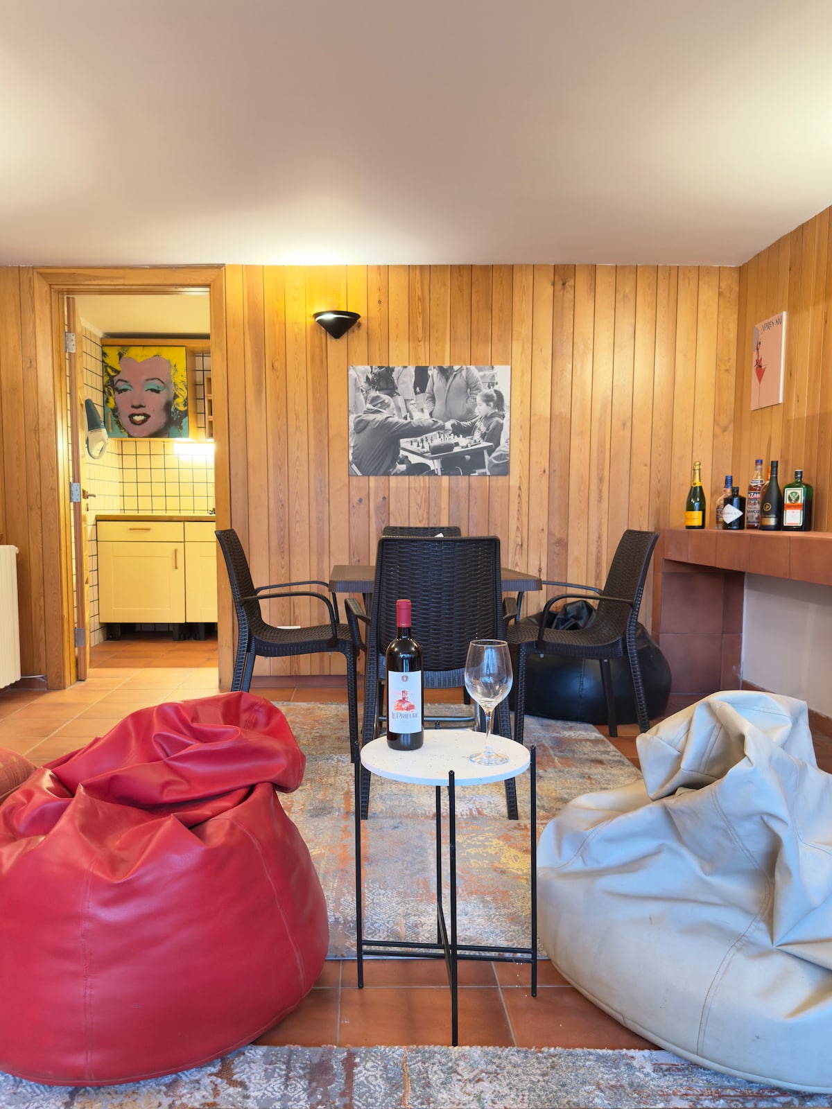A welcoming seating area features two vibrant bean bags in red and cream, complemented by a small table holding a wine glass and a bottle of wine. Wooden paneling contrasts with a light-toned rug, while a subtle light fixture illuminates the space.