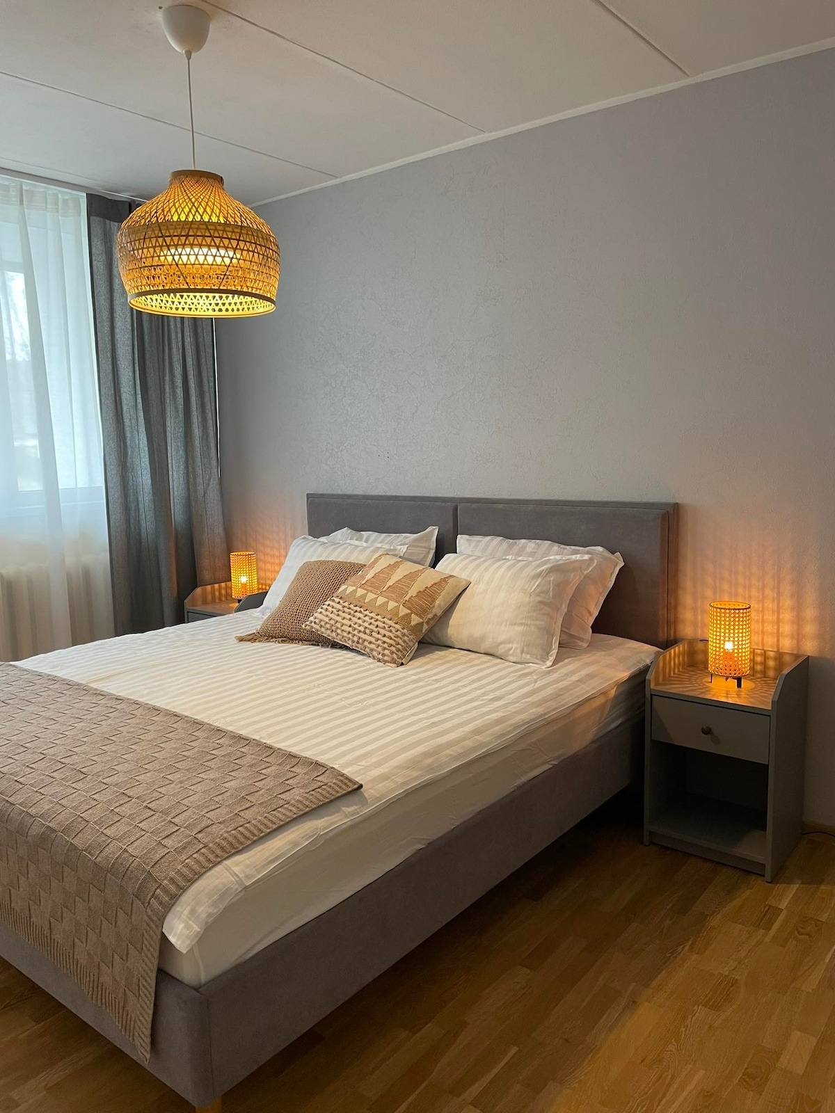 A cozy bedroom features a spacious bed with white linens and decorative pillows. Soft lamps on either side illuminate the room, complemented by a woven pendant light overhead. Light curtains allow natural light to filter in, enhancing the serene atmosphere.