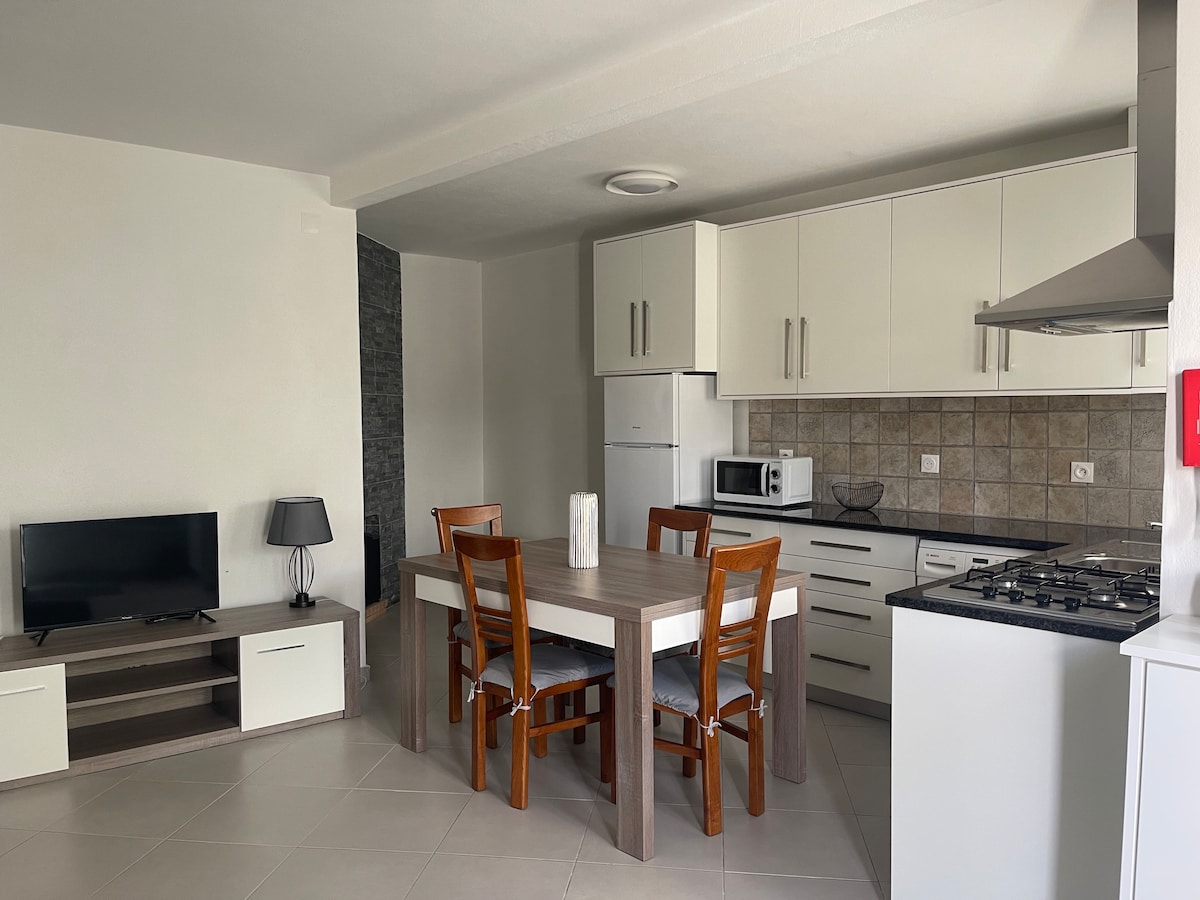 A modern kitchen area is visible, featuring a dining table with wooden chairs. A television is placed on a low cabinet, and a white refrigerator and microwave are included in the kitchen cabinetry. Light-coloured tiles enhance the open and airy atmosphere.