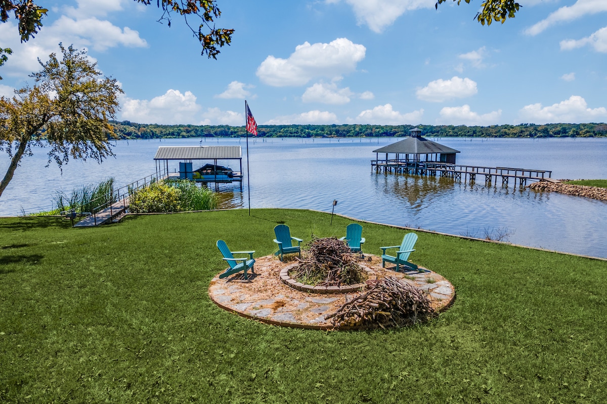 A serene outdoor area features a circular stone patio surrounded by green grass, with multiple Adirondack chairs arranged for relaxation. In the background, a private dock extends into the lake, framed by gentle waves and lush greenery under a clear blue sky.