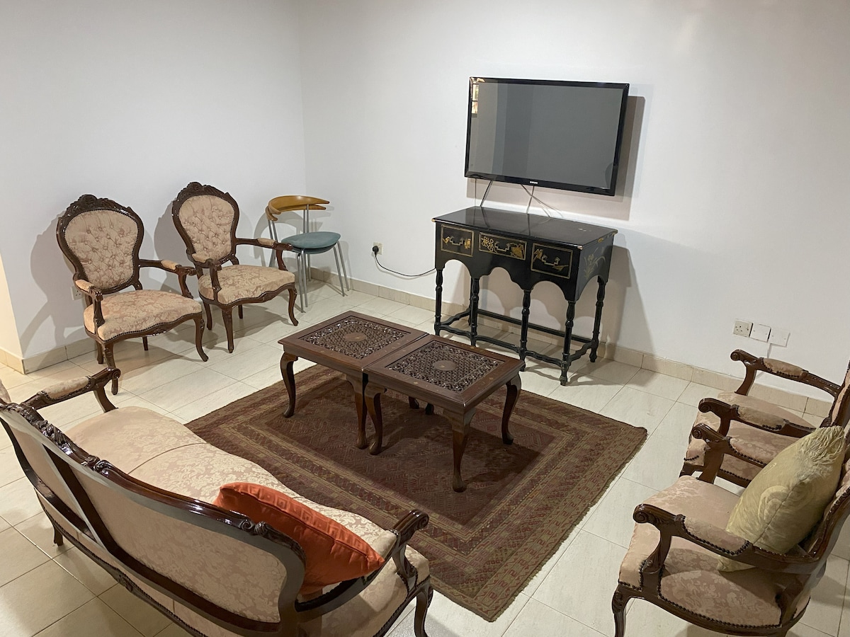 The living room showcases a cozy arrangement of antique furnishings, featuring two elegant armchairs and a long sofa, complemented by a decorative coffee table. A flat-screen television is mounted on the wall, and soft lighting enhances the serene ambiance of the space.