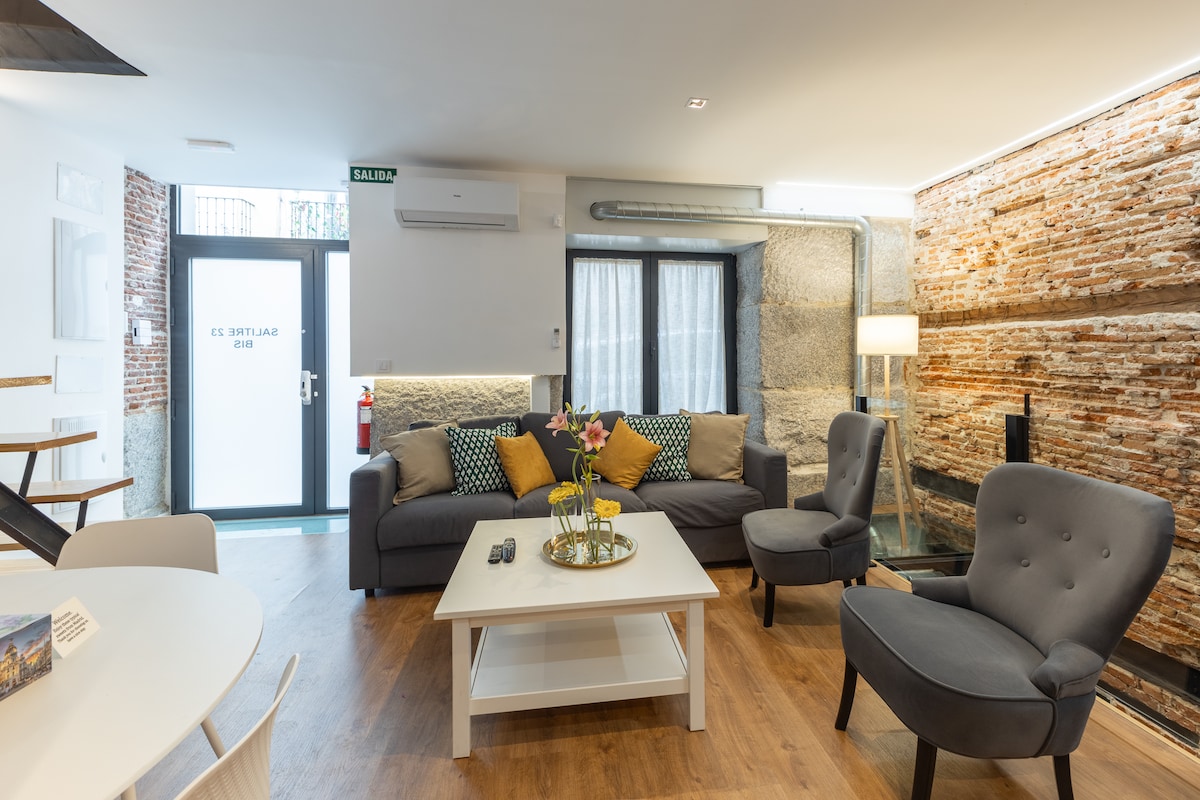 The living area features a comfortable grey sofa adorned with colorful cushions. A white coffee table sits in the center, while accent chairs offer additional seating. Large windows allow natural light to brighten the space, complemented by exposed brick walls that add character.