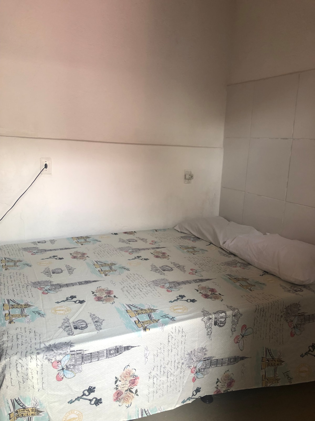 A spacious bed is covered with a patterned bedspread featuring landmarks and floral designs. A single white pillow sits against the wall. The room is neutrally painted, with light entering from an unseen source, contributing to a bright atmosphere.