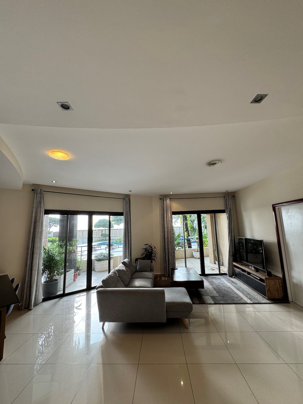 The spacious living area features large glass doors that open to a terrace, allowing natural light to fill the room. Neutral-toned furnishings, including a comfortable sofa, are complemented by a sleek television and decorative plants, creating a relaxed atmosphere.