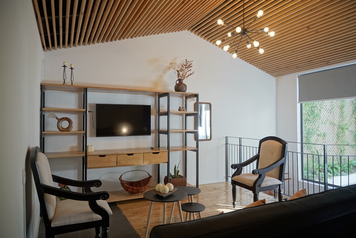 A modern living area is showcased with wooden slatted ceilings and natural light flowing through a large window. A media unit holds a flat-screen television, while seating is provided by two upholstered chairs and a comfortable sofa, accompanied by a decorative bowl and minimalistic decor.