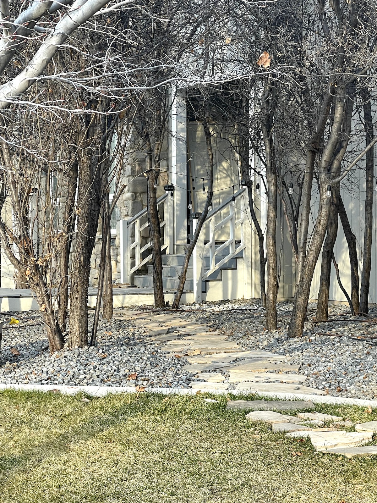 A stone pathway leads to the entrance, flanked by slender trees and gravel landscaping. The exterior door is partially visible at the top of a small flight of steps, offering a glimpse of the apartment's private entrance.