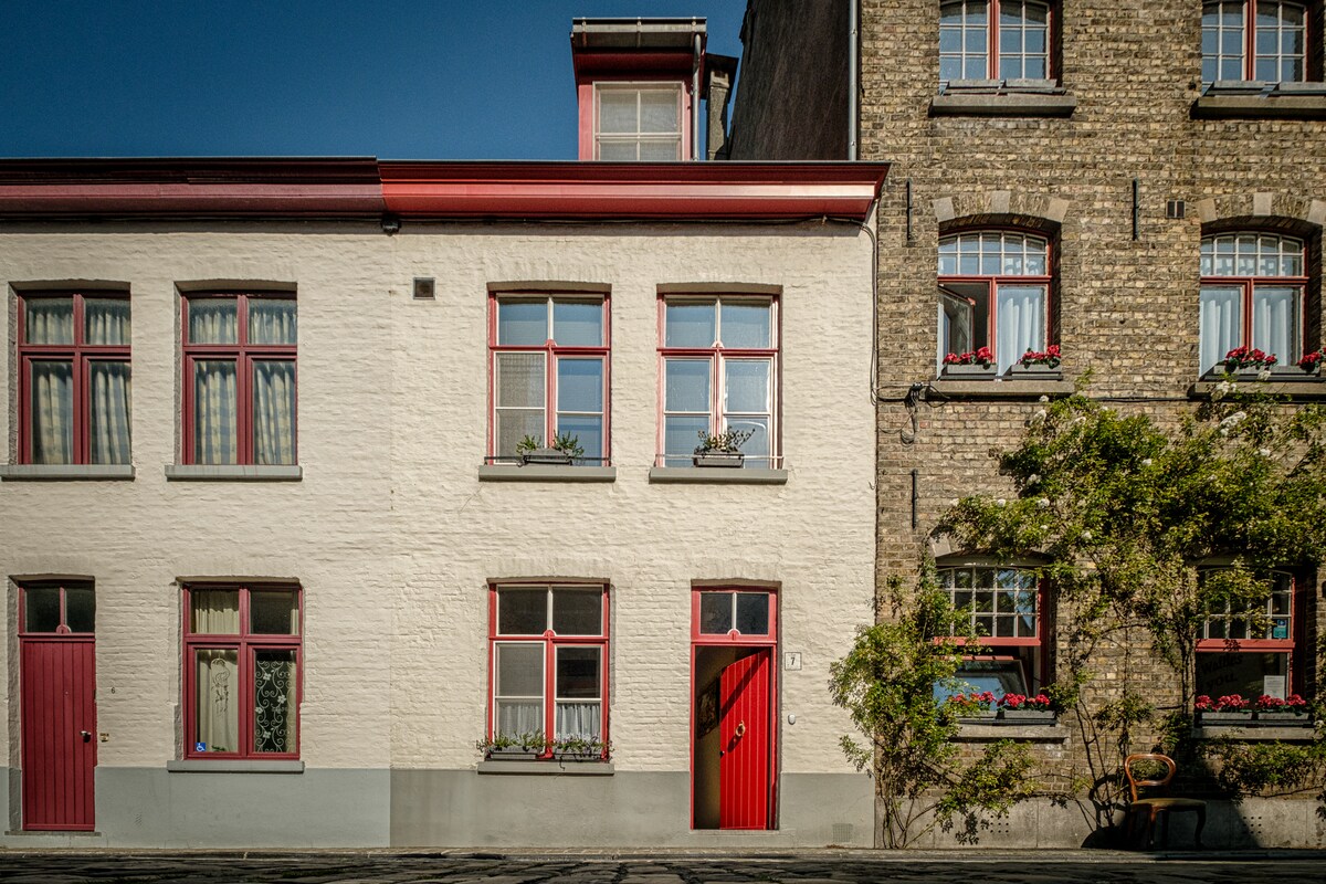 A charming two-story building is displayed with a white exterior and red accents. The windows are adorned with flower boxes, and a bright red door enhances the inviting appearance. The neighboring structure features a contrasting brick facade, contributing to the neighborhood's historic character.