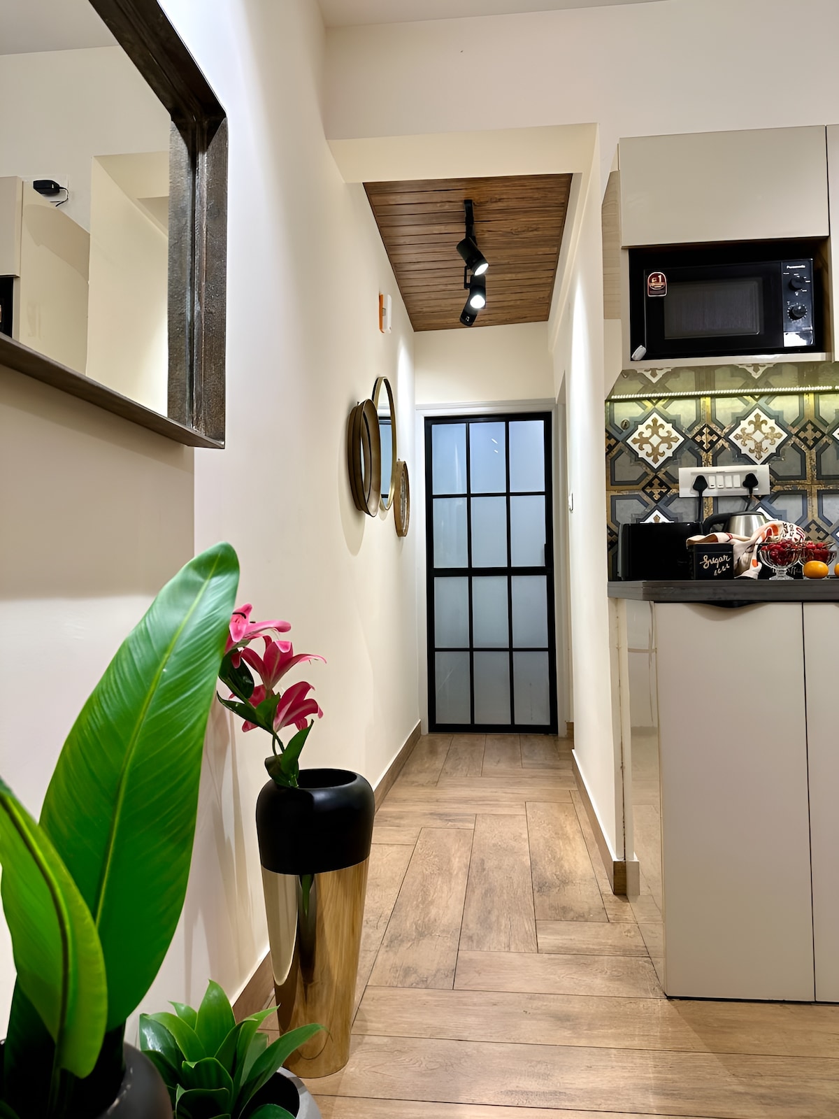 The corridor is adorned with decorative mirrors and features a tall indoor plant in a black vase. Light wood flooring contrasts with white walls, while intricate tile patterns are showcased in the kitchen area. A glimpse of a frosted glass door can be seen at the end of the hallway.