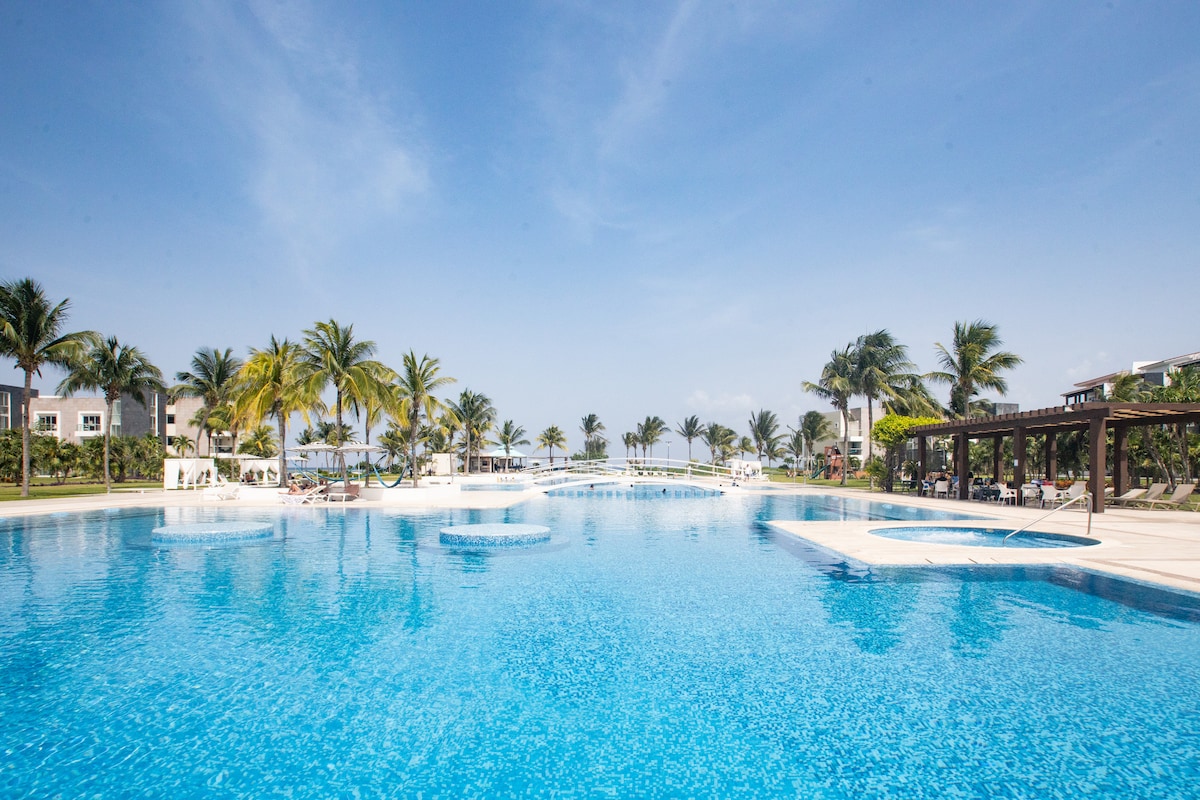 A large, inviting pool takes center stage, surrounded by vibrant palm trees. Lounge chairs are positioned along the water's edge, and a tranquil atmosphere is created by the expansive layout. The clear blue sky meets the shimmering surface of the pool, reflecting the inviting atmosphere.