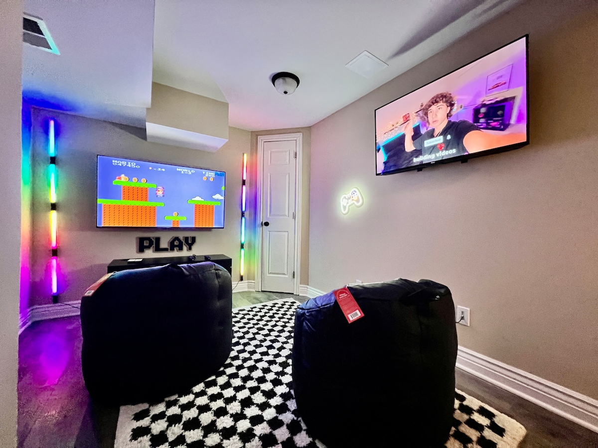 A vibrant game room features two large televisions mounted on the walls. Neon lighting enhances the playful atmosphere, and a distinct 'PLAY' sign is displayed. Comfortable bean bags are positioned on a patterned rug, inviting relaxation and enjoyment within the space.