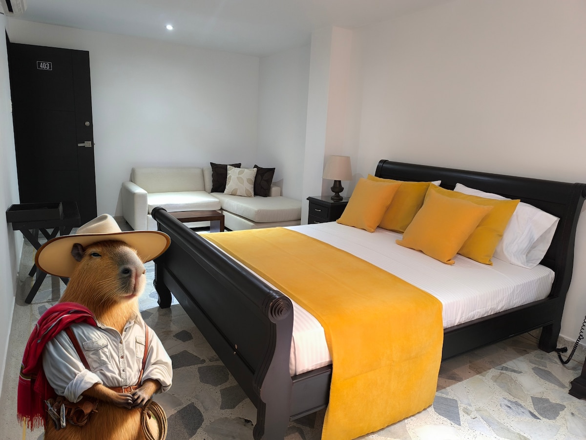 A spacious room is depicted, featuring a large bed with bright yellow accent pillows and a cozy blanket. A comfortable sofa sits across from the bed, complemented by a side table with a lamp. The overall decor is clean and minimal, with light-colored walls and stylish flooring.