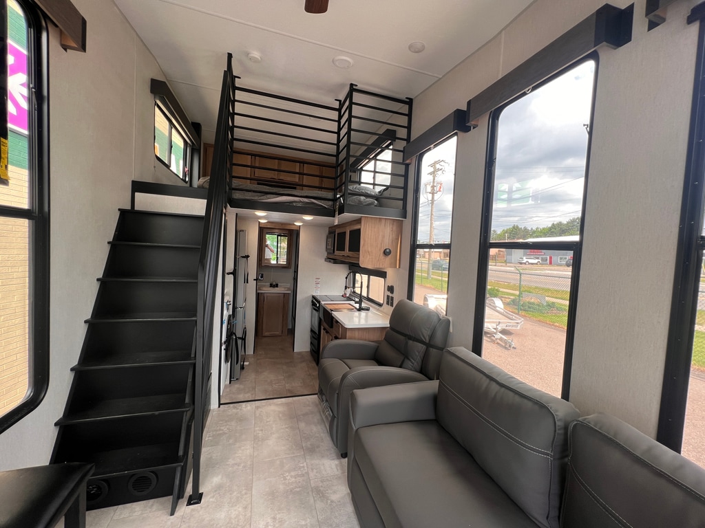 An open living area features a sleek gray leather sofa positioned near large windows, providing views of the outdoors. A spiral staircase leads to a lofted bedroom. The kitchen, equipped with modern appliances, is visible in the background, enhancing the functional layout of the space.