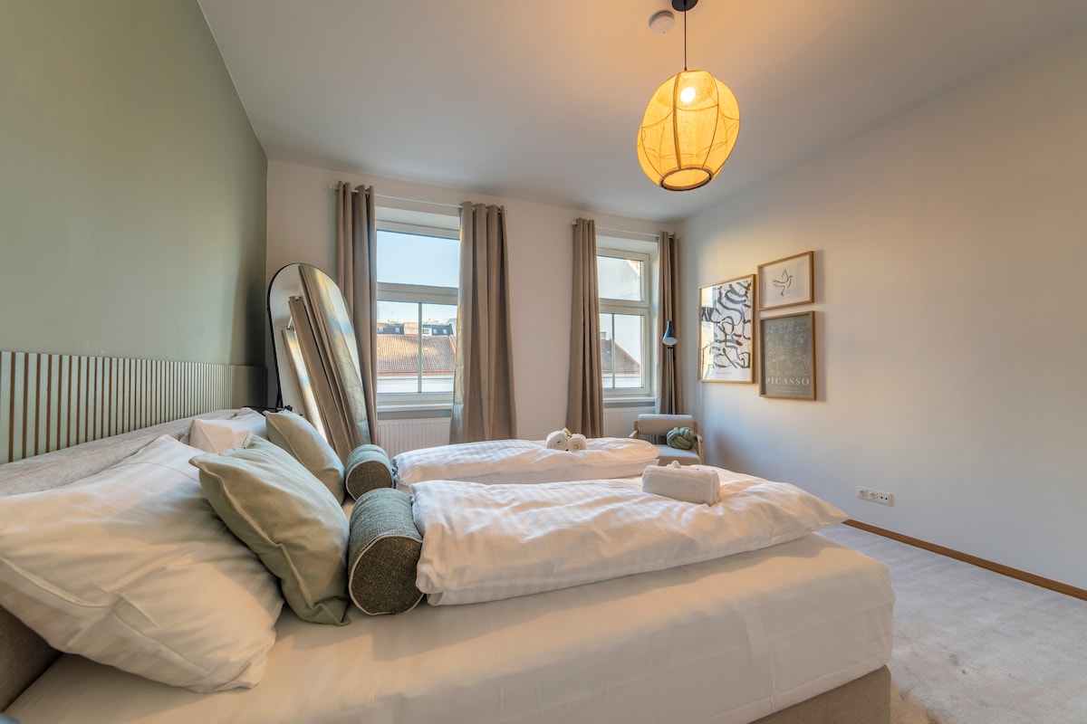 A well-lit bedroom includes two spacious beds positioned side by side, accented with decorative pillows. A large mirror reflects natural light from the windows, which are draped with soft curtains. Artistic wall decor is displayed, contributing to a calming atmosphere.
