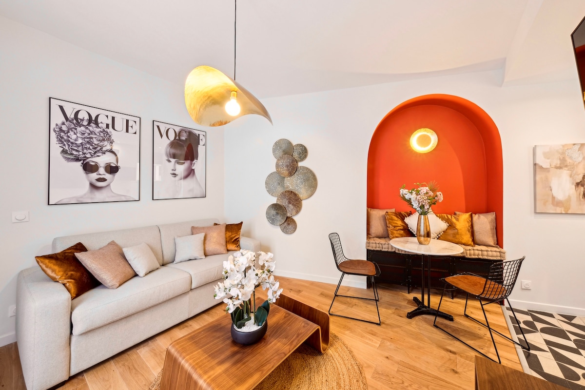 The living area features a light-colored sofa complemented by decorative pillows. A round dining table with stylish black chairs is positioned beside an accent wall in warm red. Fashion-themed artwork adorns the walls, and a contemporary pendant light hangs overhead.