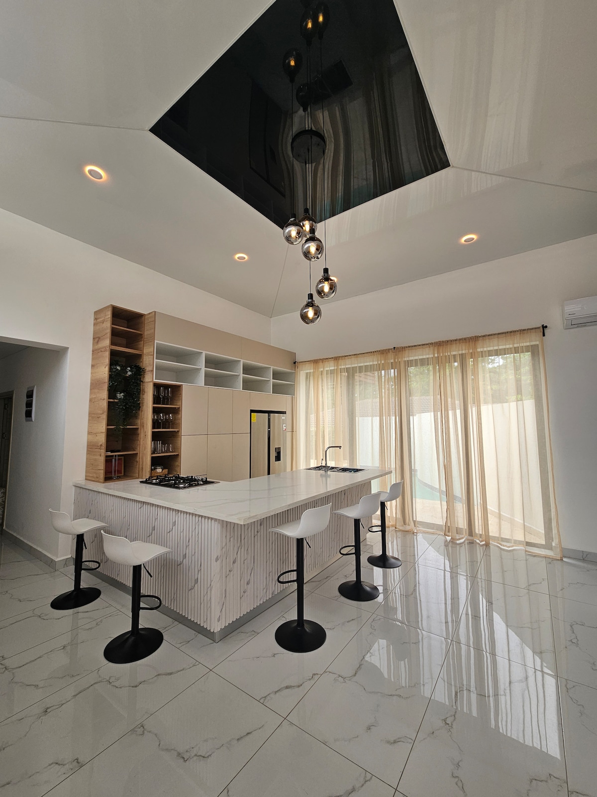 A modern kitchen is featured with a sleek island and four high stools. Large windows allow natural light to fill the space, complemented by soft curtains. The stylish ceiling showcases unique lighting fixtures, adding a contemporary touch to the overall design.