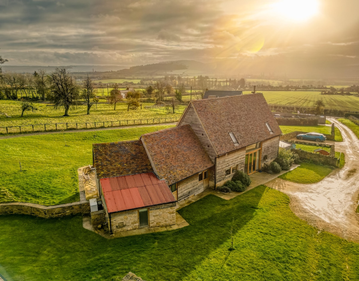 The barn is set in a serene countryside landscape, featuring a blend of rustic stone and wooden architecture. A vibrant green lawn surrounds the structure, and sweeping fields extend into the distance under a soft, golden sunset.