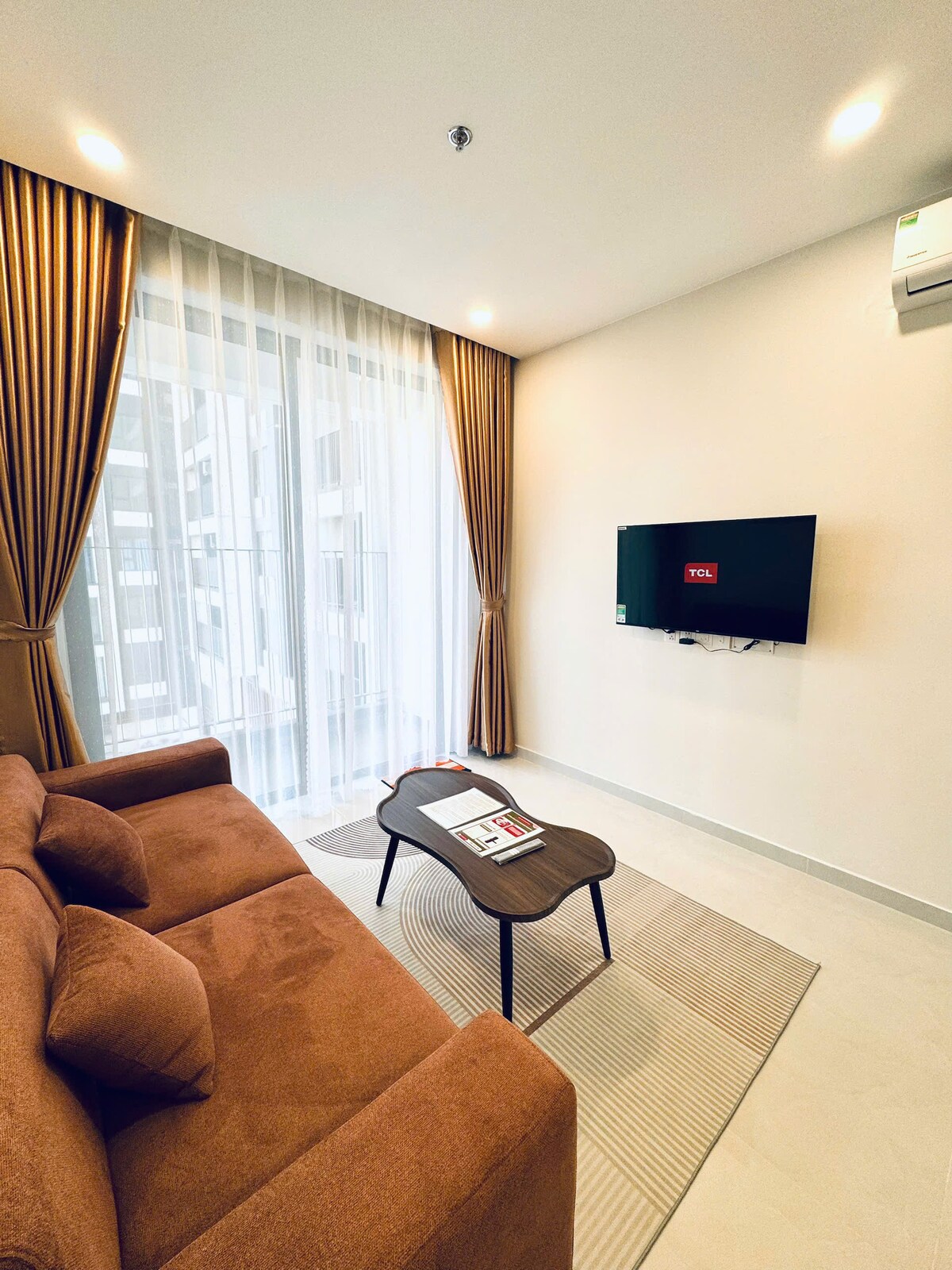 A modern living area features a comfortable brown sofa positioned opposite a mounted television. Large windows adorned with light drapes allow ample natural light to brighten the space. A small coffee table sits on a neatly arranged rug, providing a welcoming spot for relaxation.