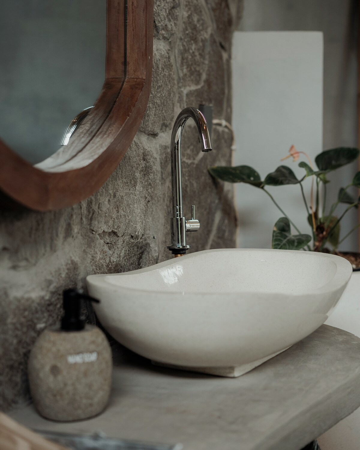 A natural stone sink is placed atop a smooth countertop, complemented by a sleek silver faucet. The rustic stone wall provides a textured backdrop, while a circular wooden mirror above the sink enhances the organic feel. A small potted plant adds a touch of greenery.