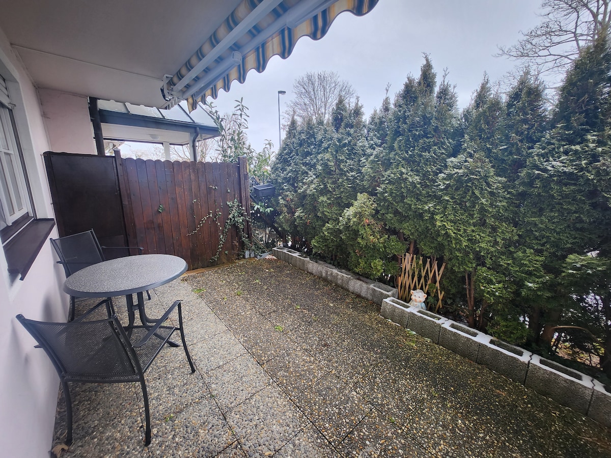 An outdoor terrace is displayed, featuring a round table accompanied by two chairs. A sturdy protective awning extends overhead. Lush greenery borders the space, providing a sense of privacy. The ground is made of stone tiles, creating a functional and open area for relaxation.