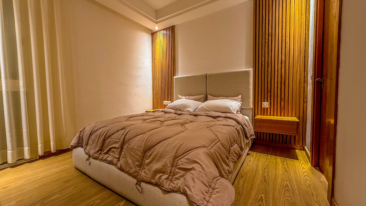 A spacious sleeping area features warm wooden accents and soft lighting. The bed is dressed with a neutral-toned comforter and several pillows. Sheer curtains frame a window, allowing natural light to gently illuminate the room, contributing to a serene ambiance.