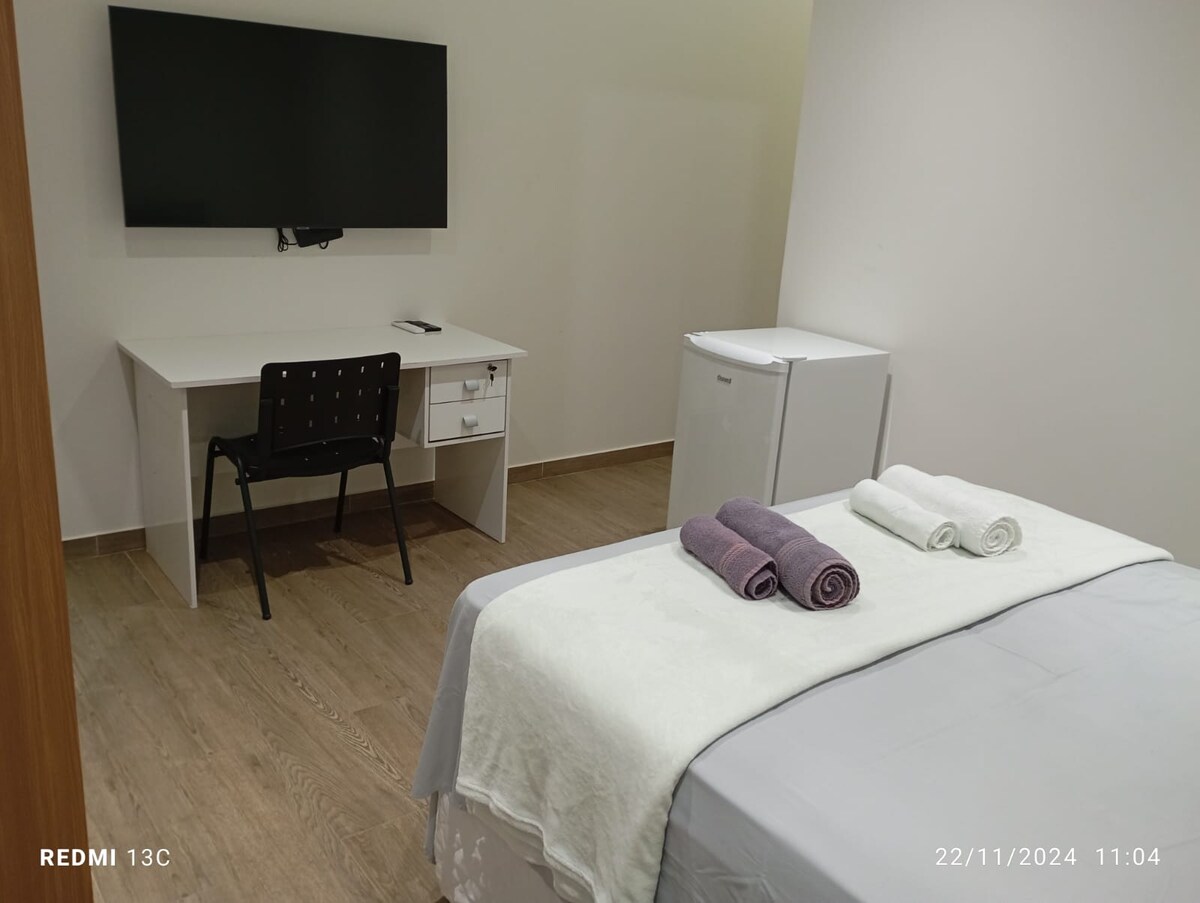 A comfortable sleeping area features a spacious bed with neatly arranged towels. A desk with a chair is positioned nearby, alongside a compact refrigerator. A flat-screen TV is mounted on the wall, contributing to the room's functionality and modern design.