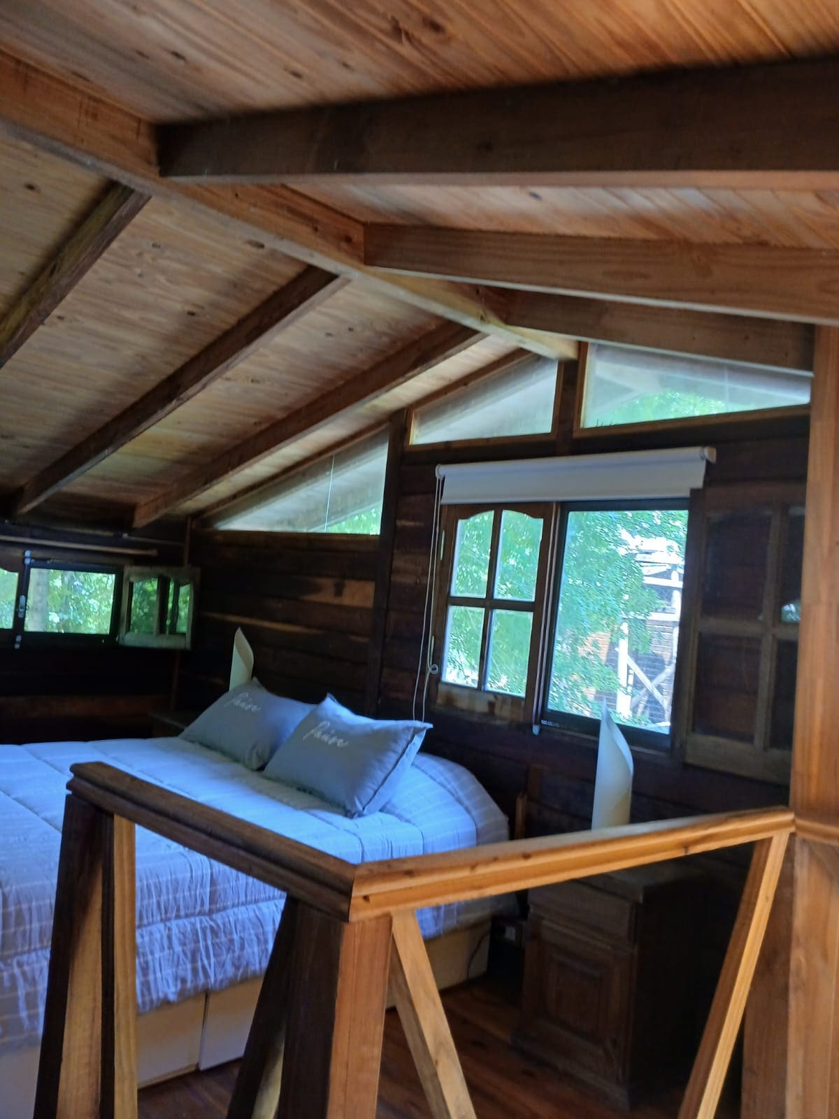 A spacious sleeping area features a large bed adorned with soft, muted pillows. Natural light filters through large windows framed in wood, highlighting the rustic character of the room. The ceiling showcases exposed wooden beams, creating a cozy, inviting environment.