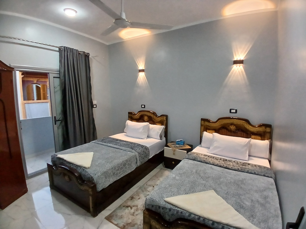 A serene guest room features two wooden-framed beds adorned with soft linens. A decorative rug rests beneath, and walls are softly illuminated by stylish wall sconces. A nightstand holds small items, and curtains frame a nearby window, allowing natural light to enter the space.