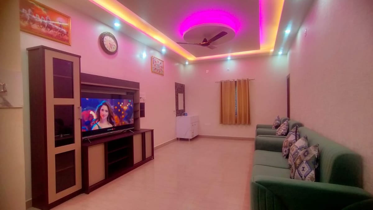 A bright living area is adorned with colorful LED lights along the ceiling. A dark wood entertainment unit houses a large television, while a white dresser offers additional storage. Comfortable green sofas, complemented by patterned cushions, are arranged to create an inviting space.