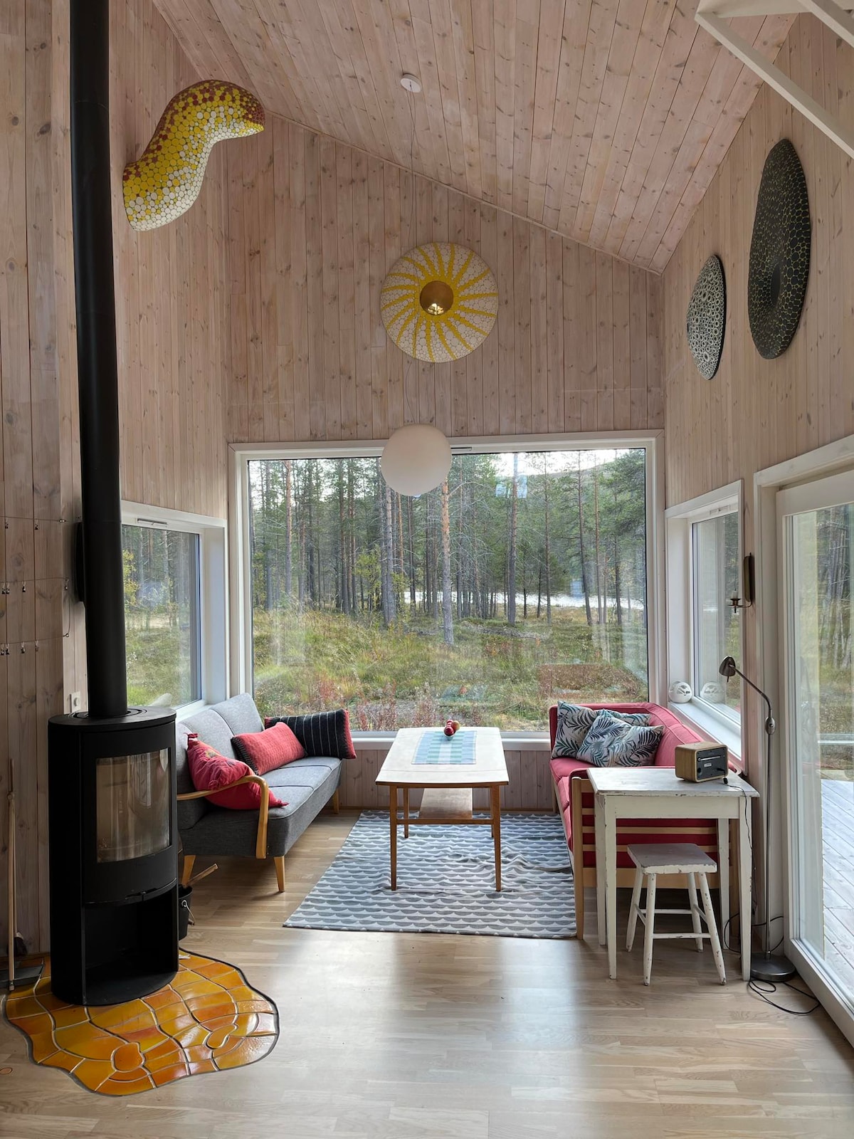 The bright living area is surrounded by large windows offering views of the forest. A modern wood stove stands prominently, while a comfortable seating arrangement invites relaxation. Unique wall decorations add character, and a dining table is positioned near the cozy seating.
