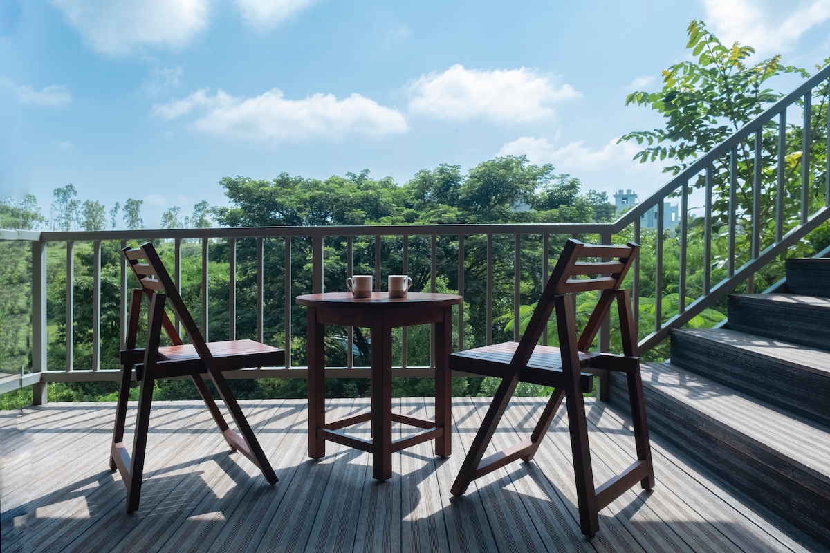 A balcony area is presented with two wooden chairs and a small round table, set against a backdrop of lush greenery. Bright blue skies and soft clouds enhance the tranquil outdoor space, inviting relaxation.
