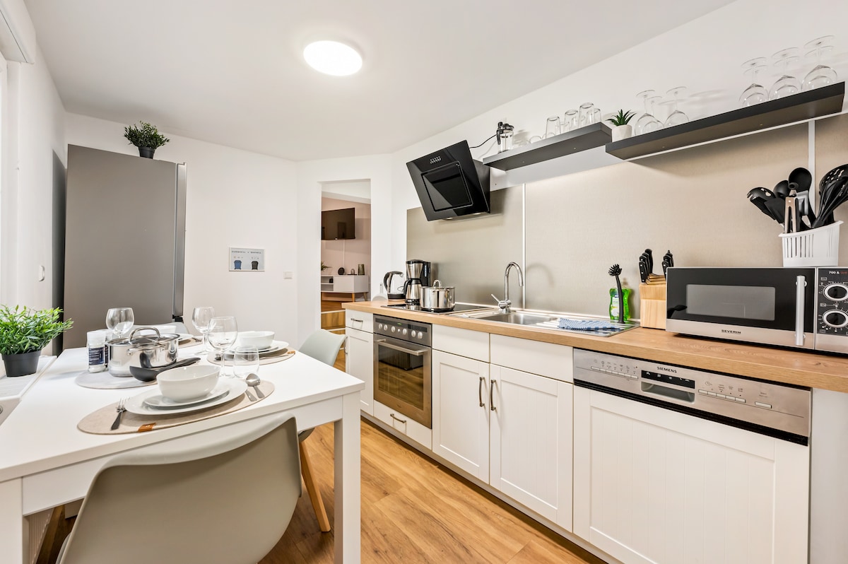 A bright and fully equipped kitchen features a modern design with wooden countertops and white cabinetry. The space includes essential appliances such as a stove, oven, microwave, and a coffee machine. A dining table is set, ready for a meal, enhancing the functional layout.