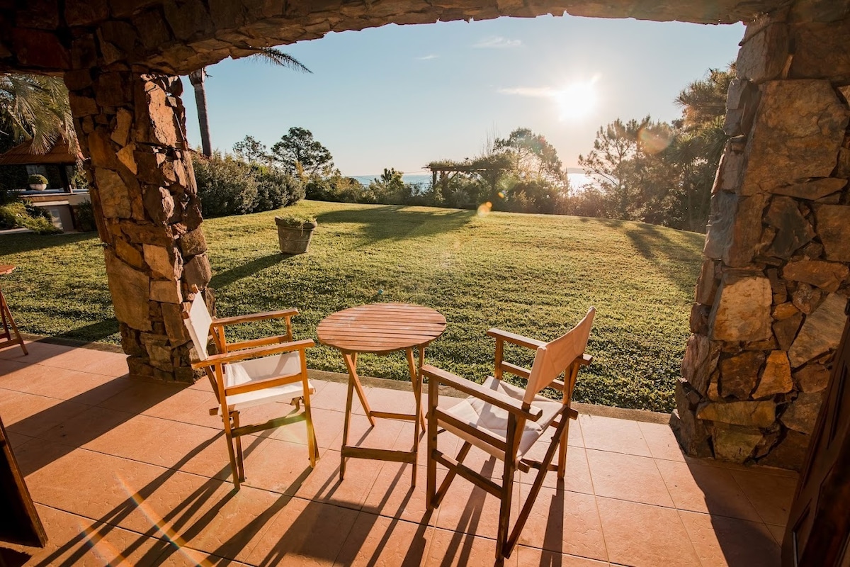 An outdoor seating area features two wooden chairs and a small round table positioned on a tiled patio. The surrounding green lawn extends toward a backdrop of trees, with the sun setting on the horizon, casting a warm glow across the scene.
