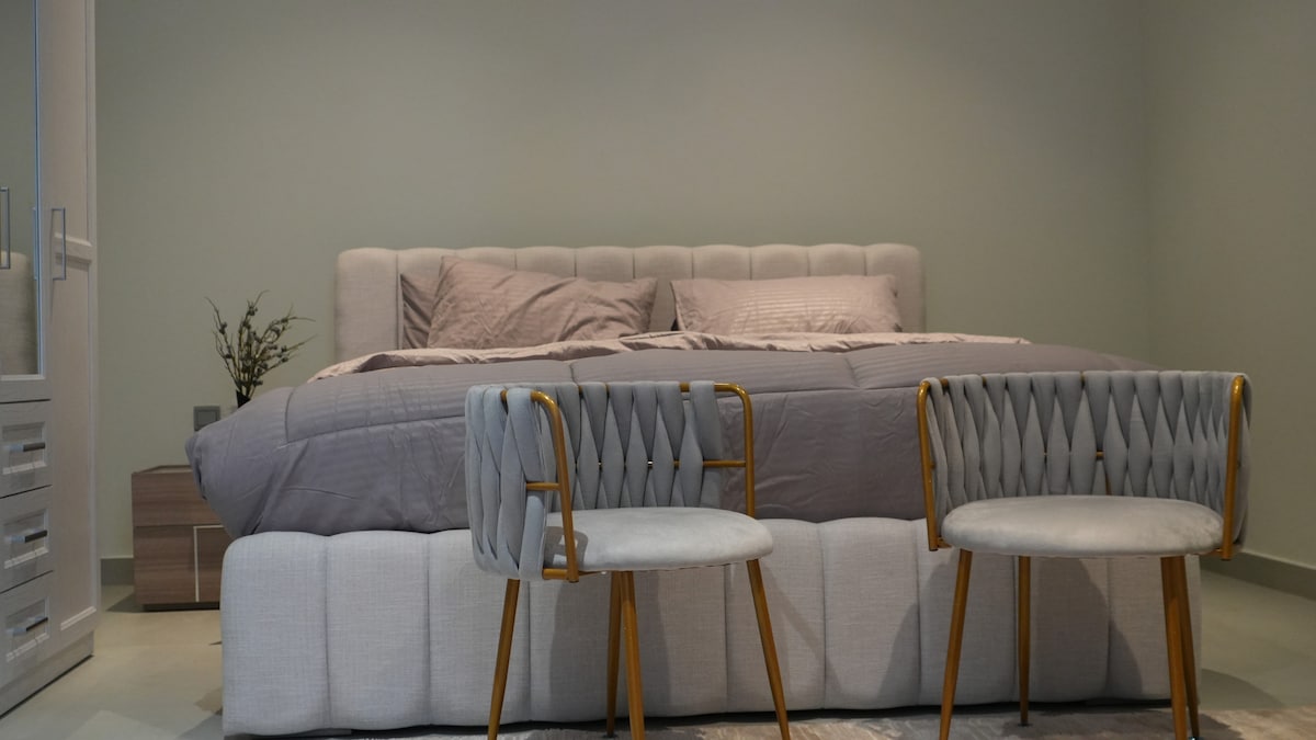 A spacious sleeping area features a comfortably dressed bed with a neutral-toned coverlet and a simple headboard. Two stylish chairs with gold accents are positioned in front of the bed, creating a cozy and inviting seating arrangement.
