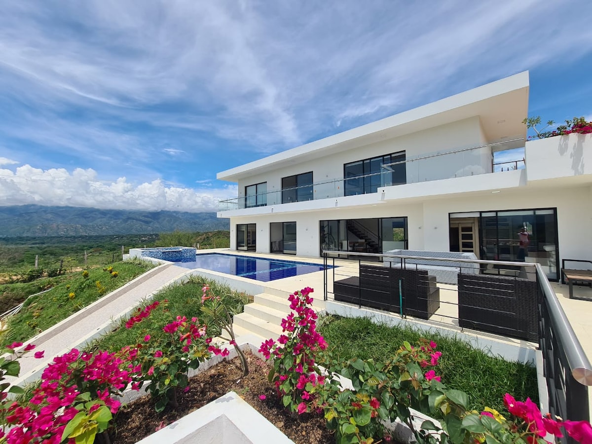 A modern villa is showcased against a backdrop of rolling green hills and a clear blue sky. A spacious pool reflects the sky and is surrounded by a terrace. Vibrant pink flowers enhance the outdoor area, framing the path leading to the entrance.