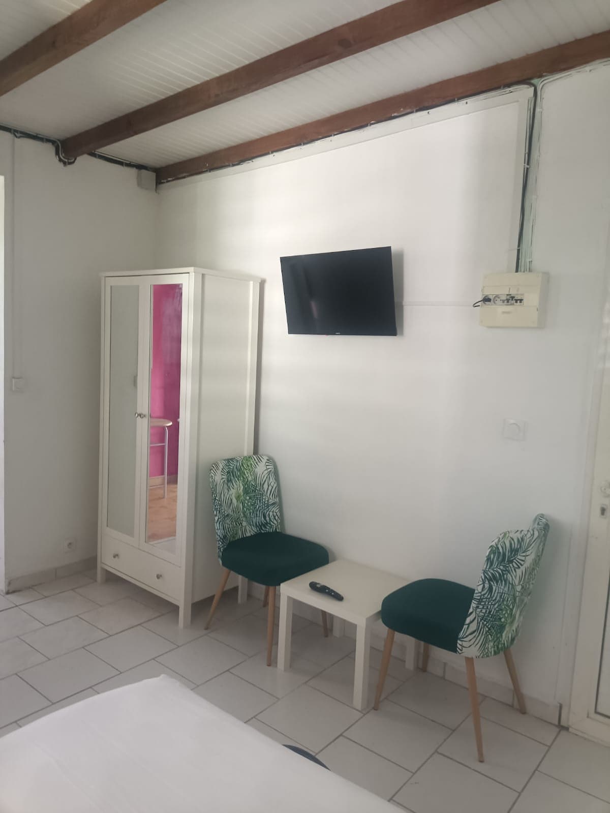 A bright living area features two chairs with green tropical-patterned upholstery and a small table. A sleek white wardrobe stands against the wall, and a wall-mounted television is positioned for viewing. White walls and tiled flooring create a clean, open space.
