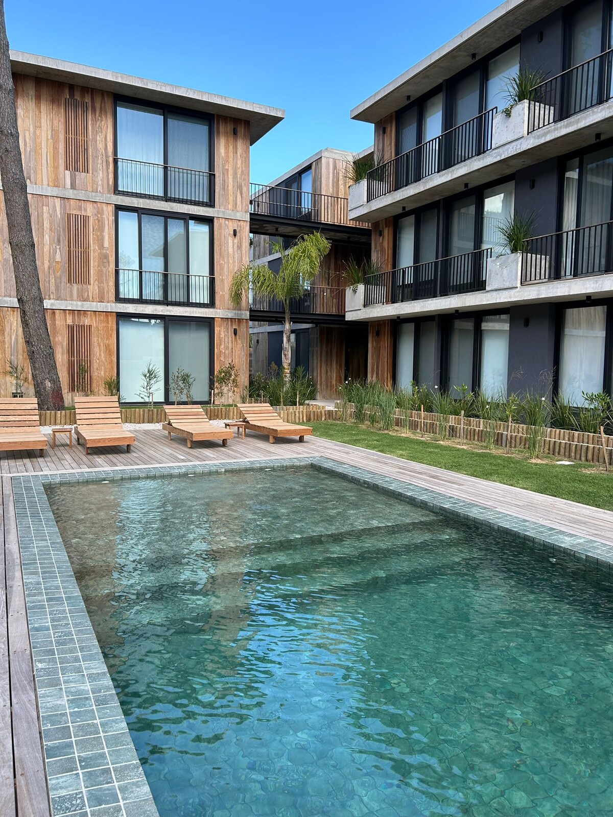 A serene outdoor pool is framed by wooden deck chairs, positioned beside a clear water feature. The surrounding architecture includes modern, multi-story buildings with large glass windows and balconies, complemented by lush greenery that enhances the tranquil environment.