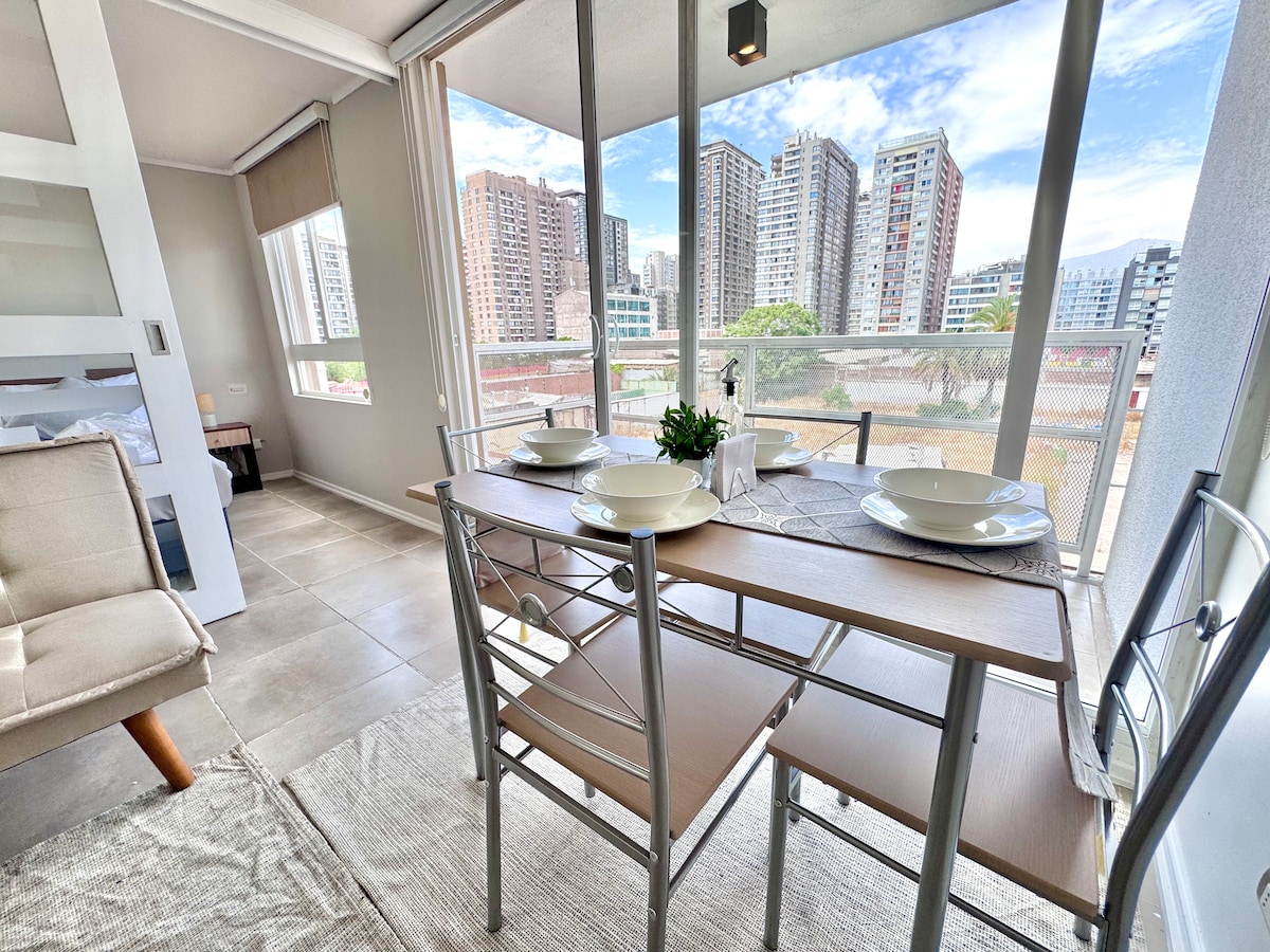 A dining area features a light wooden table set for two, accompanied by silver metal chairs. Large glass doors open to a balcony with views of surrounding buildings and the city skyline, allowing natural light to fill the space.