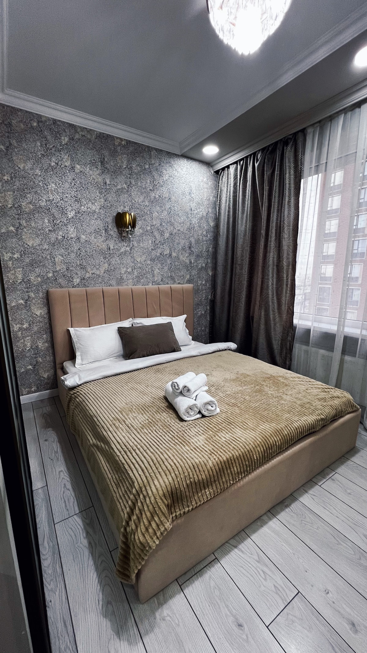 A serene sleeping area features a spacious bed with a textured beige blanket. A subtle accent pillow is placed against the headboard. Natural light filters through sheer curtains, illuminating the elegant wallpaper and enhancing the cozy atmosphere. A ceiling light fixture adds soft illumination.