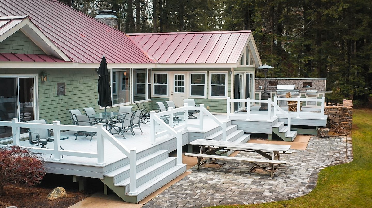 A spacious deck area is highlighted, featuring a picnic table and outdoor dining set with several chairs. The green exterior of the house complements the red metal roof. A barbeque grill is visible nearby, with stone pathways and a serene wooded backdrop.