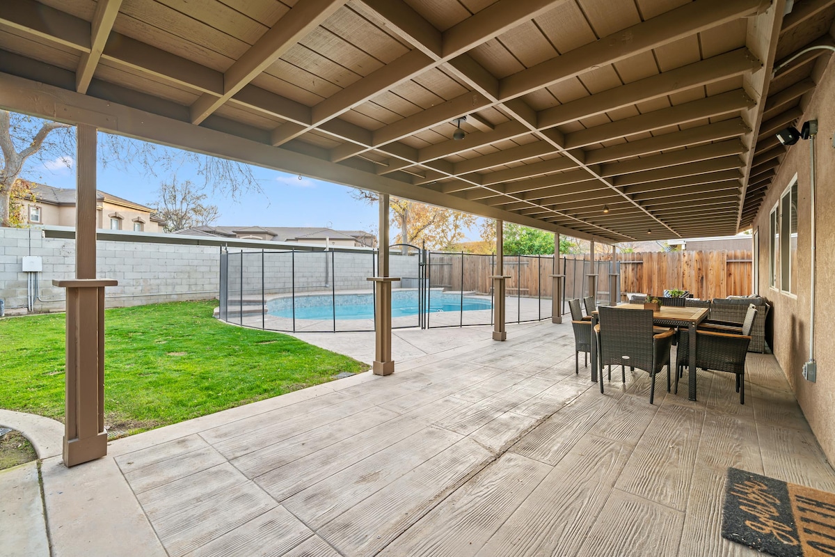 An outdoor covered patio is presented, featuring a dining area with chairs and a table. A spacious green yard is visible behind a secure fence, with a swimming pool reflecting the blue sky. The concrete flooring provides ample space for relaxation.