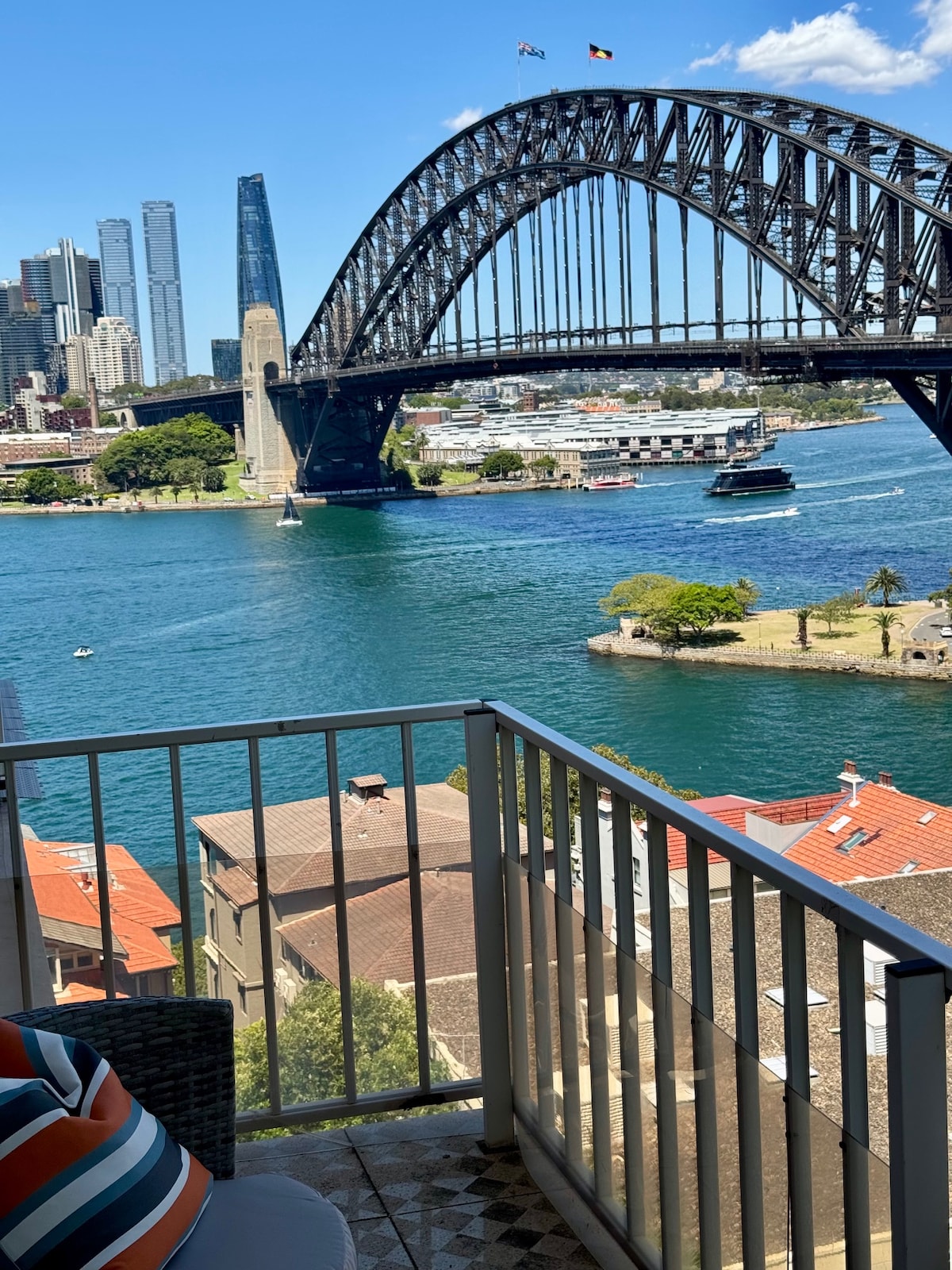 A scenic view of the Sydney Harbour Bridge is captured from a balcony, showcasing the vibrant blue waters below. Modern skyscrapers stand in the background, with boats passing under the bridge. Lush greenery and local architecture are visible in the foreground, creating a lively atmosphere.