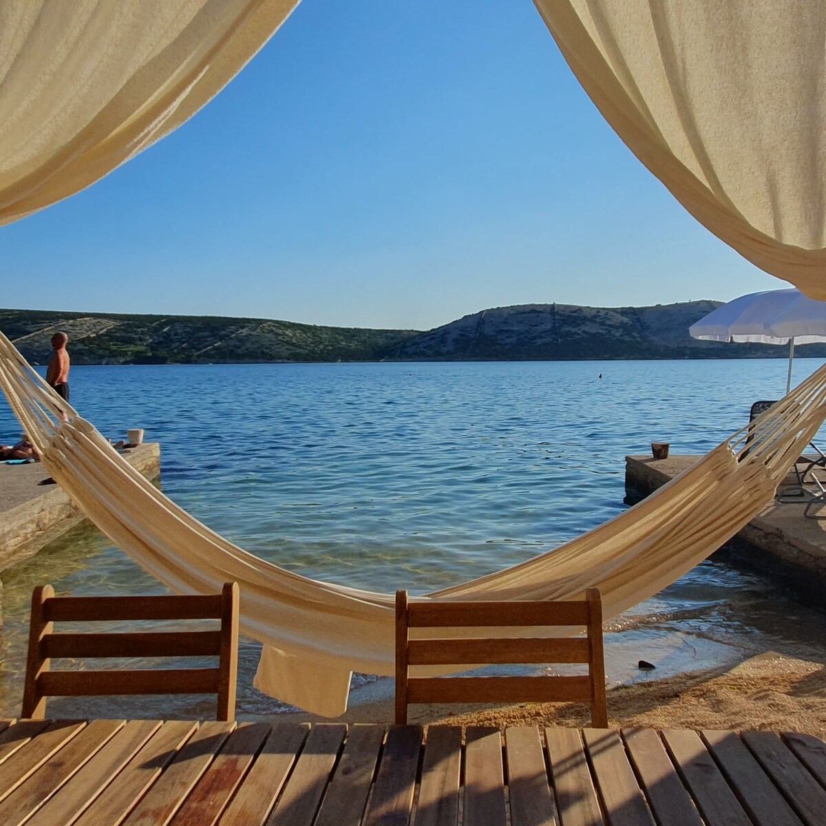 A serene view of the calm water is framed by two hammocks draped between wooden supports, creating a relaxed atmosphere. Gentle waves lap at the shore, with green hills visible in the background under a clear blue sky. Sun loungers rest nearby, inviting relaxation.