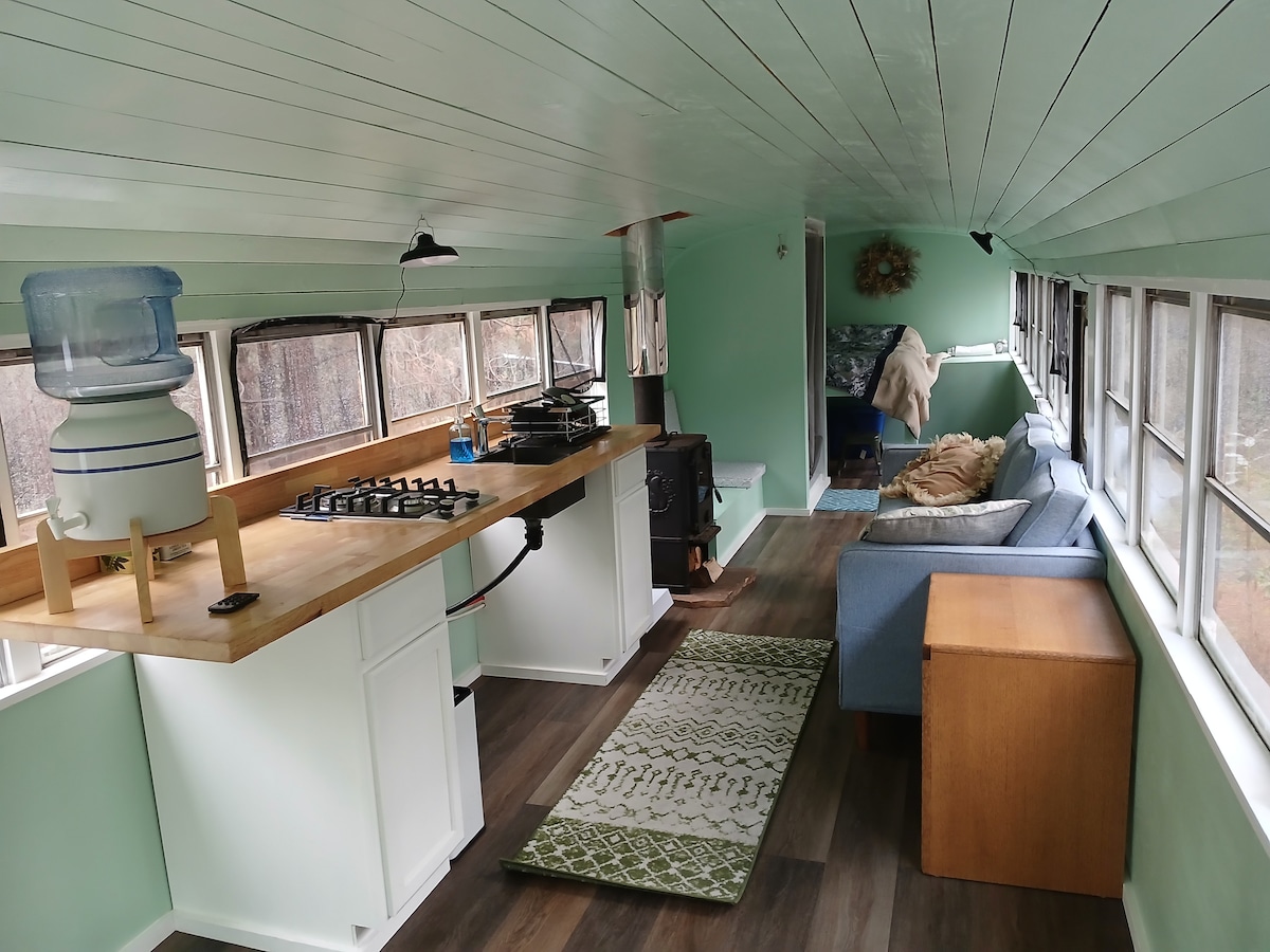 An open interior features light green walls and a high ceiling. A wooden countertop with a gas stove is positioned against one wall, while comfortable seating is arranged along another. Large windows provide ample natural light, enhancing the serene atmosphere.